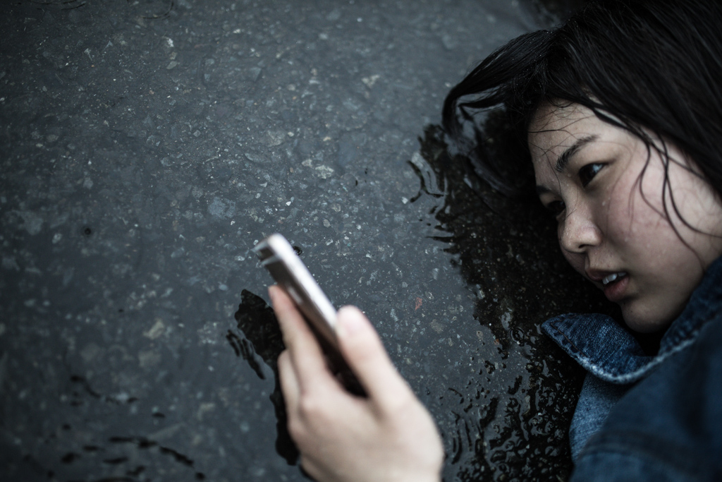 悲しそうに雨に打たれながらアスファルトに寝転がりスマホを見つめる女性