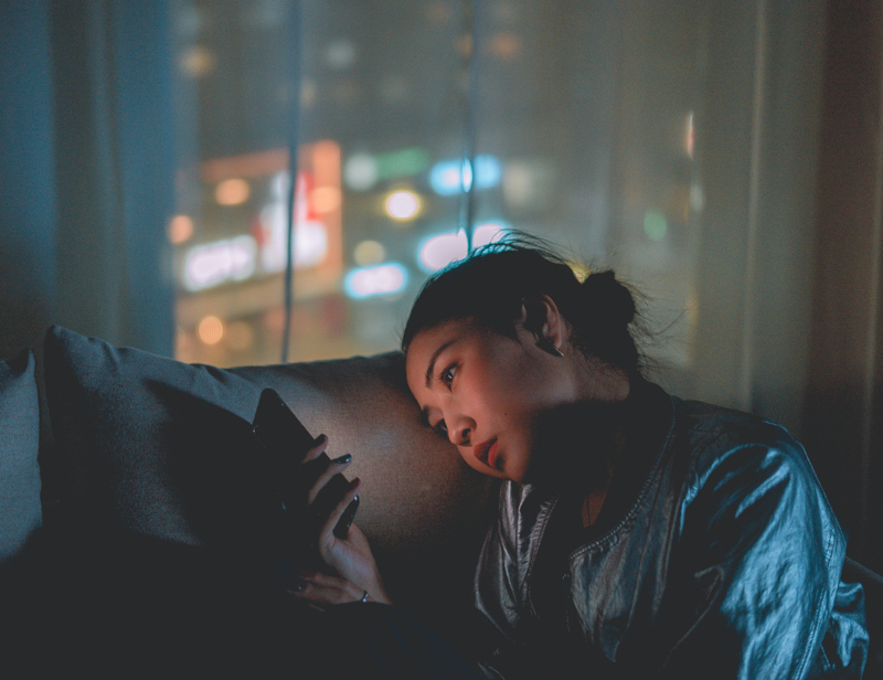 夜の部屋のソファで項垂れながらスマホをいじる女性
