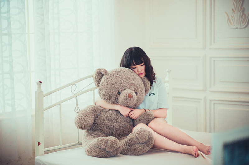 ベッドの上で大きな熊のぬいぐるみを抱きしめて寂しそうにする女性